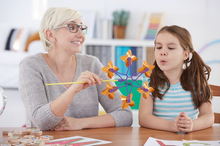 A girl is doing a blowing exercise during a speech therapy session, with the speech therapist sitting next to her